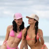 Two women in patterned bikinis and trucker hats stand on a beach, smiling and looking at each other against a blurred seaside background.