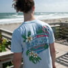 A person with long hair in a ponytail stands on a wooden boardwalk near the beach, wearing a light blue Ron Jon Surf Shop T-shirt with a colorful graphic on the back.