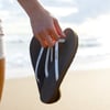A person holds a pair of black flip-flops with white straps while standing on a sandy beach near the water.