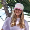 Woman smiling outdoors, wearing a light pink cap and lavender shirt. Tropical foliage in the background.