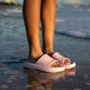 Close-up of a person standing on wet sand at the beach, wearing light pink slide sandals and an anklet, with waves in the background.