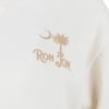 Close-up of a off white fabric with a brown graphic featuring a crescent moon, a palm tree, and the text RON JON.