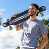 A man in a grey t-shirt smiles while holding a skateboard over his shoulder outdoors, with palm trees and a blue sky in the background.