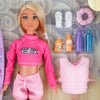 A doll with long blonde hair wearing a pink crop top and shorts, accompanied by accessories like flip-flops, hair tie, life vest, and bottles.