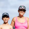 A woman and a boy, both wearing baseball caps, smile outdoors under a blue sky. The woman wears a pink top; the boy is shirtless.