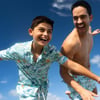 A smiling boy and man in matching floral beachwear joyfully run under a clear blue sky.
