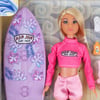 Blonde-haired doll in pink surf attire with a purple surfboard featuring floral patterns and the Ron Jon Surf Shop logo. Background includes wave designs and small accessories.
