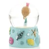 A snow globe with a small figurine of a girl in a blue dress and pink crown inside. The base is decorated with sea-themed motifs like shells and starfish.