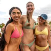 Three women in bikinis smiling outdoors, with one wearing a red checkered bikini, another in green, and the third in yellow with a blue cap. Cloudy sky and greenery in the background.