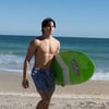 A young man wearing blue swim trunks walks on the beach holding a green Ron Jon surfboard. The ocean and sky are visible in the background.