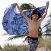 A smiling child wearing sunglasses holds up a blue beach towel with seashell designs, standing near sand and palm trees.