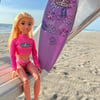 A blonde doll in a pink surf outfit sits on a metal bench at the beach next to a pink surfboard decorated with floral patterns.