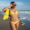 Person in a yellow bikini holding sunglasses and a beach towel, walking along a sandy beach with waves in the background.