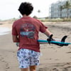 A person in a red Ron Jon Surf Shop shirt and palm tree shorts walks on the beach carrying a blue surfboard under their arm.