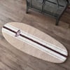 A surfboard-shaped beige, cream and brown striped rug with a RON JON SURF SHOP logo is placed on a wooden floor next to a wood burning stove.