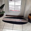 Surfboard shaped rug with black, brown, and white stripes placed in front of a glass door, surrounded by potted plants on a tiled floor.