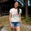 Person wearing a graphic t-shirt and denim shorts, standing outdoors near trees and a wooden shelter, smiling.