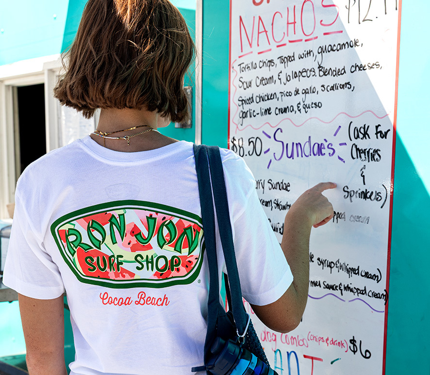 Person wearing a Ron Jon Surf Shop Cocoa Beach t-shirt points at a menu board outside a food stand painted teal.