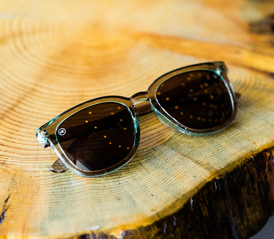A pair of sunglasses with dark lenses and clear frames rests on a cut wooden log with visible growth rings.