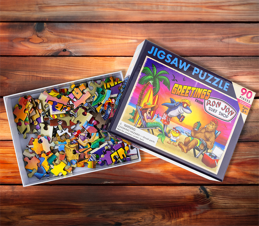 An open jigsaw puzzle box with pieces inside and a lid showing cartoon animals at a beach with text Greetings from Ron Jon Surf Shop..