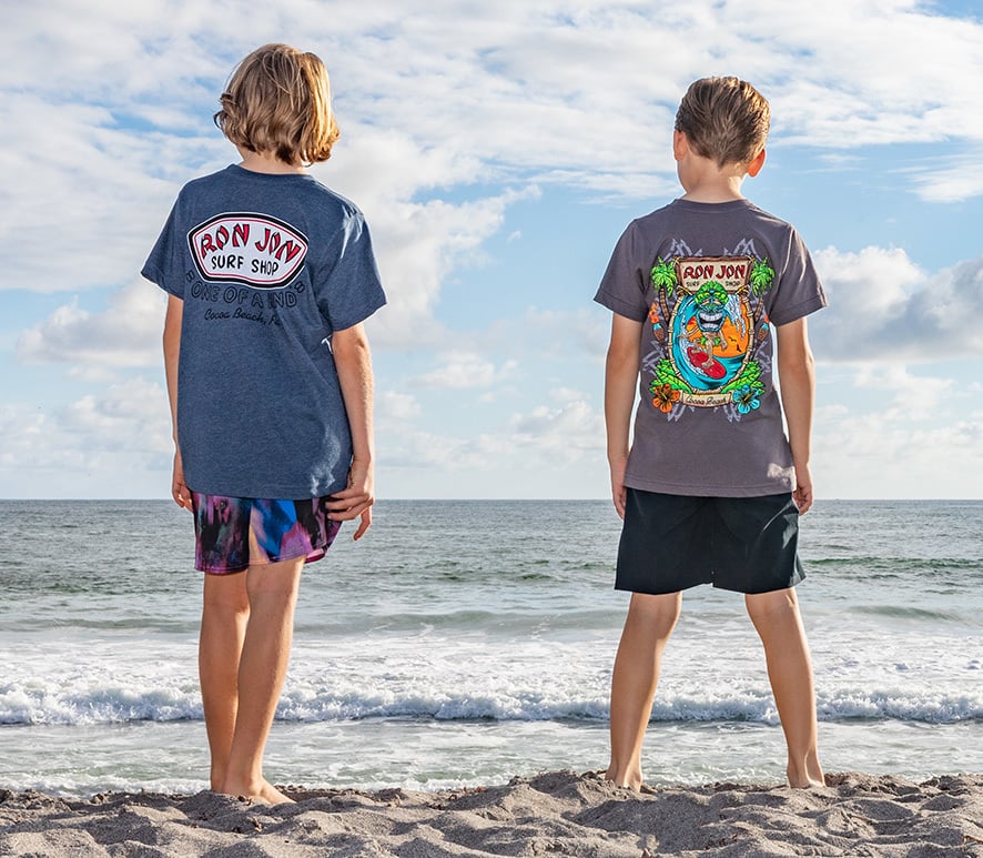 Two boys stand on a sandy beach facing the ocean, both wearing colorful Ron Jon Surf Shop t-shirts and shorts, under a partly cloudy sky.