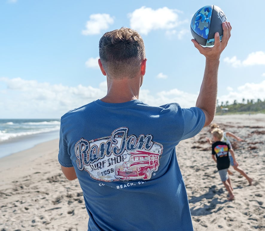A man wearing a blue Ron Jon Surf Shop T-shirt prepares to throw a football on a sandy beach, while two children run in the background near the shoreline.