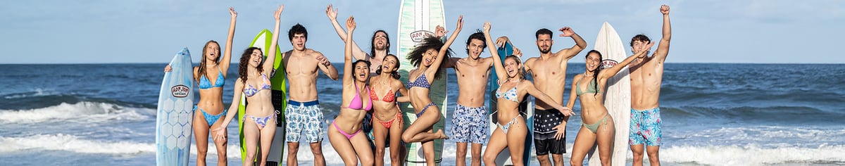 A group of people in swimsuits and board shorts stand on the beach holding surfboards, smiling and raising their arms, with waves and the ocean in the background.