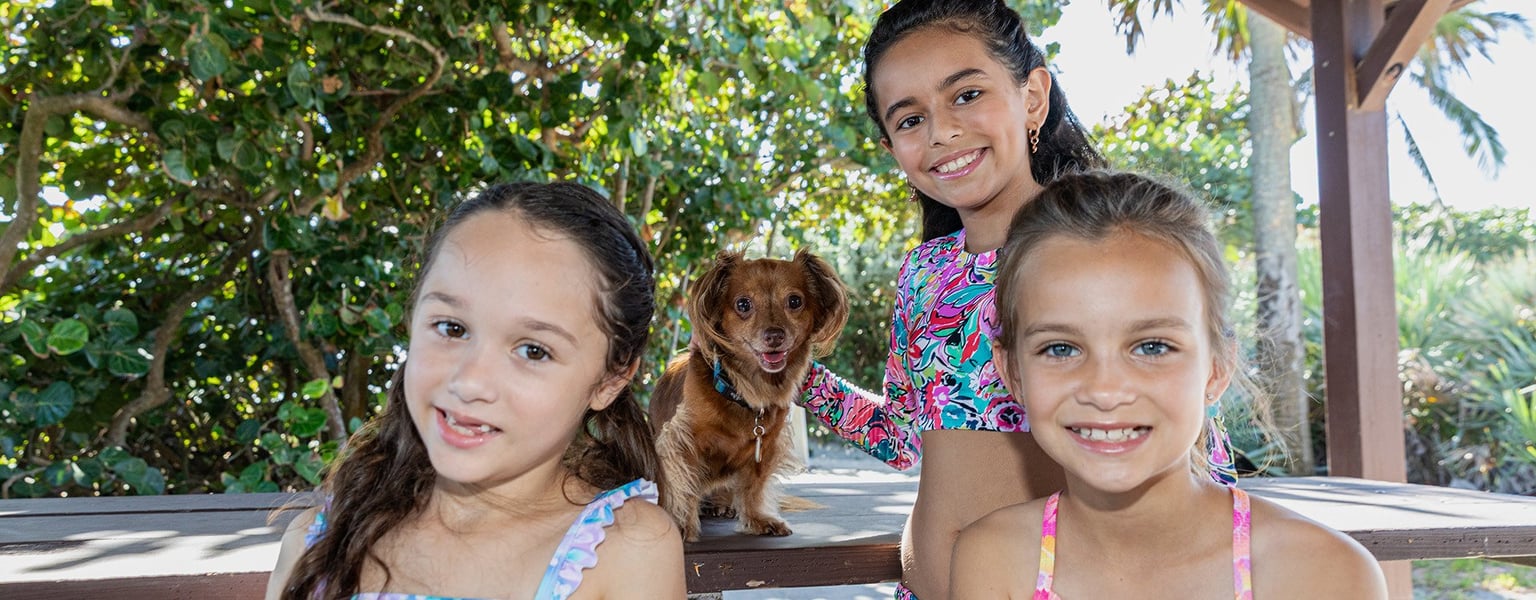 Three young girls and a small brown dog pose together outdoors under a shaded area with trees in the background.