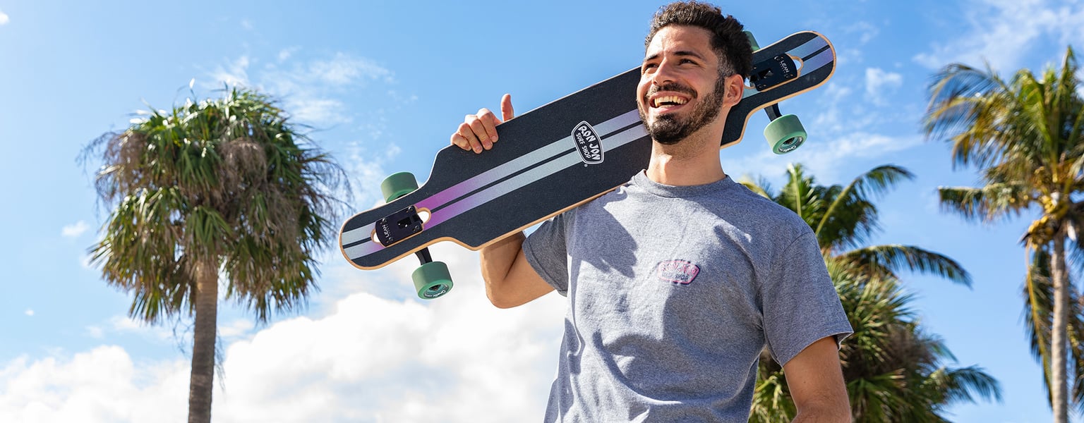 A man in a grey t-shirt smiles while holding a skateboard over his shoulder outdoors, with palm trees and a blue sky in the background.
