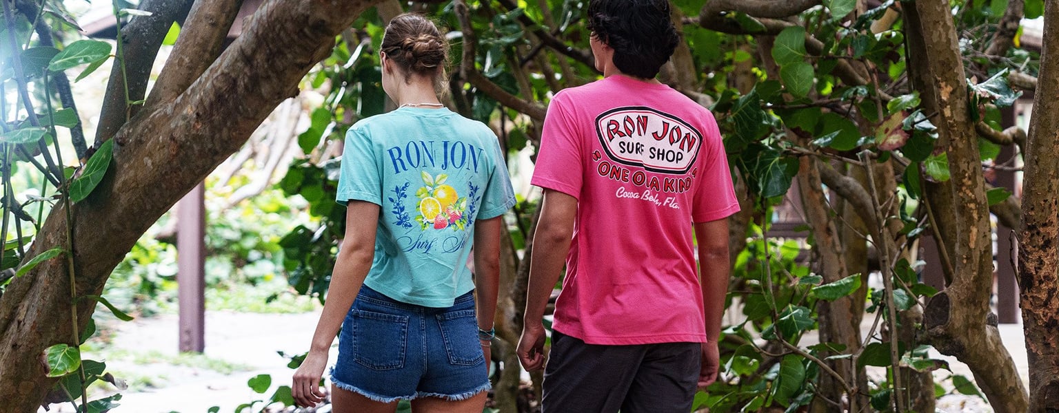 Two people walk outdoors among trees, both wearing casual shorts and Ron Jon Surf Shop t-shirts, viewed from behind.