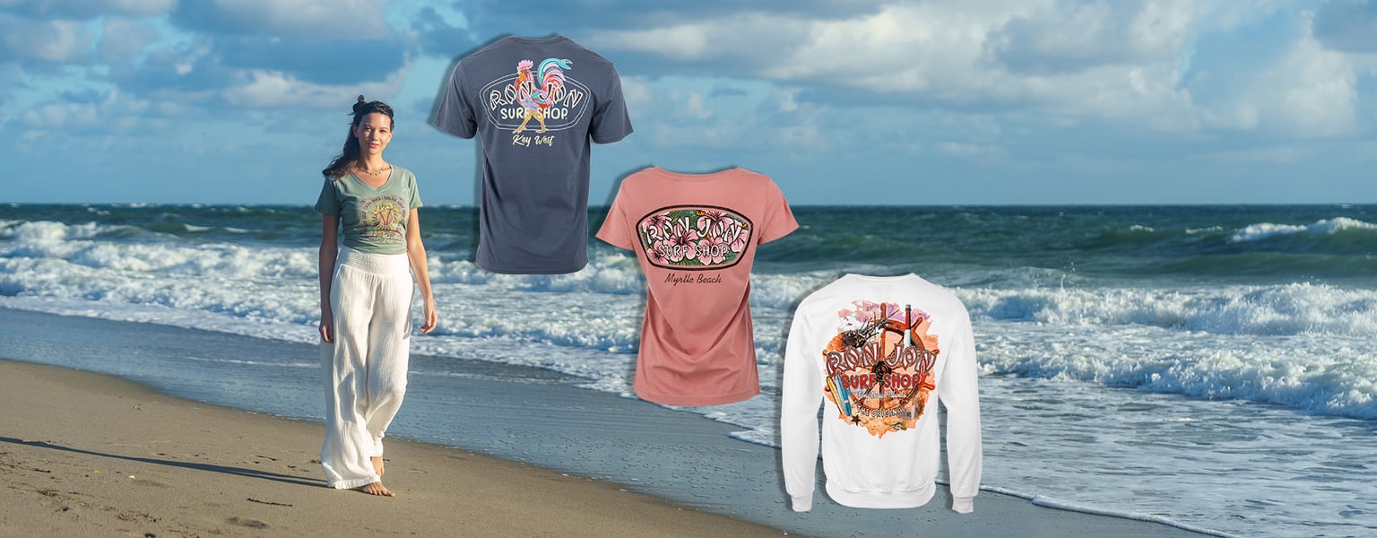 A woman walks on a beach near the waves; colorful graphic t-shirts and a sweatshirt are displayed next to her against the ocean background.