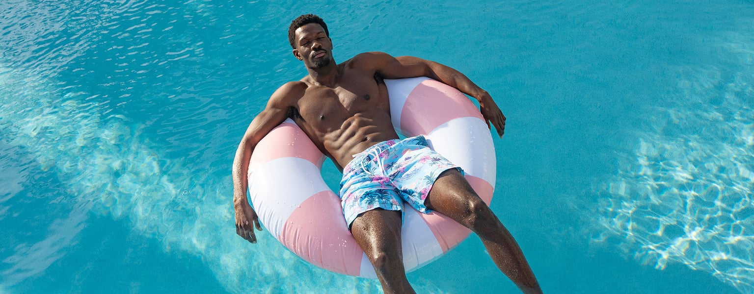 Man wearing patterned swim trunks floats on a pink and white inflatable ring in a bright blue swimming pool.