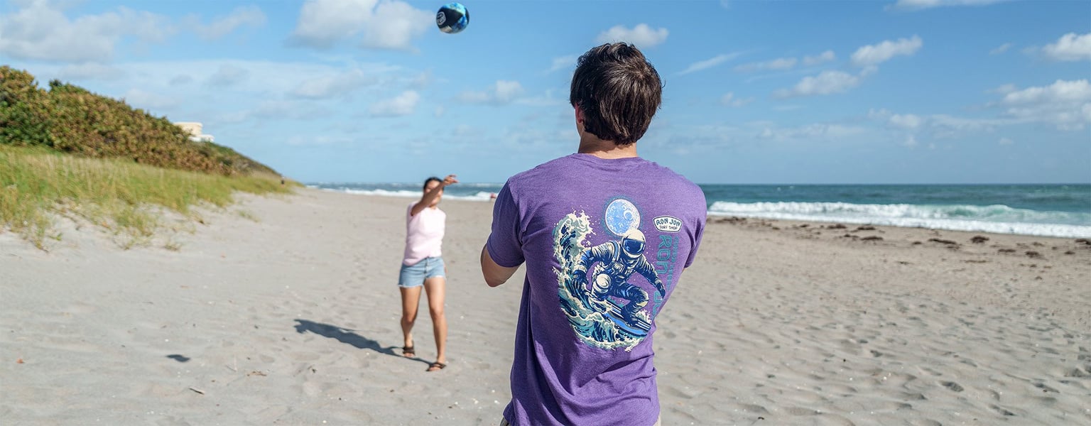 Two people play catch with a football on a sandy beach near the ocean, with clear skies and scattered clouds overhead.