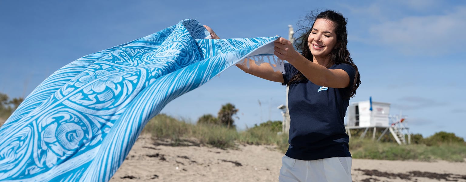 Woman standing on a sandy beach, smiling while holding and shaking out a blue patterned towel; lifeguard stand and grassy dunes in the background.