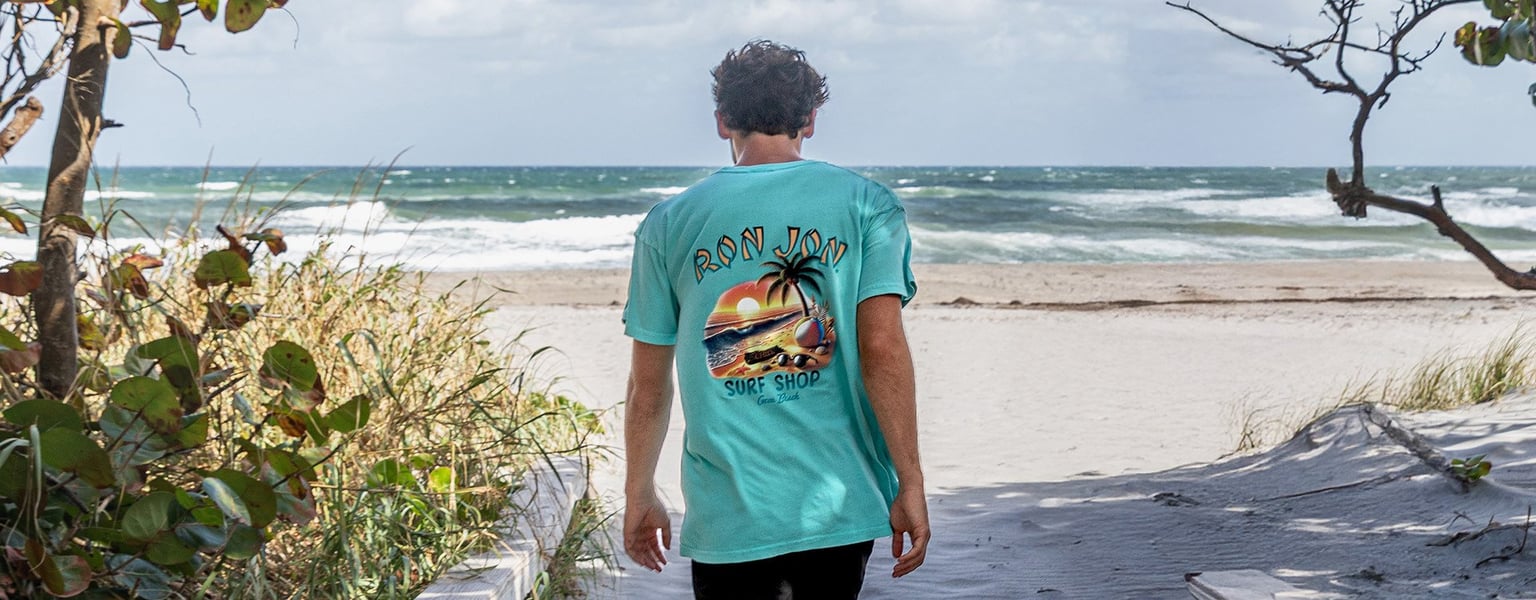 A person wearing a light blue Ron Jon Surf Shop T-shirt walks on a sandy path toward the beach with waves and cloudy skies in the background.
