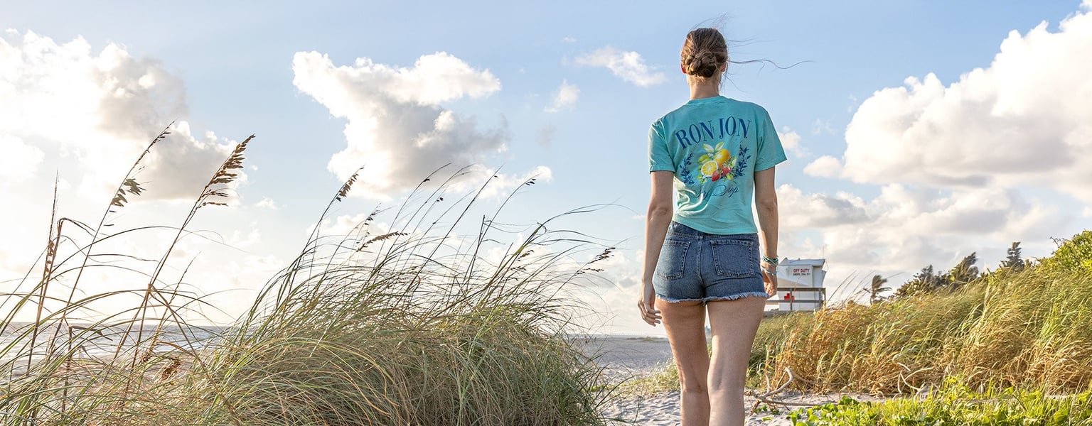 A person in a blue T-shirt and denim shorts walks along a sandy beach path with tall grass, facing the ocean and a lifeguard tower in the distance.