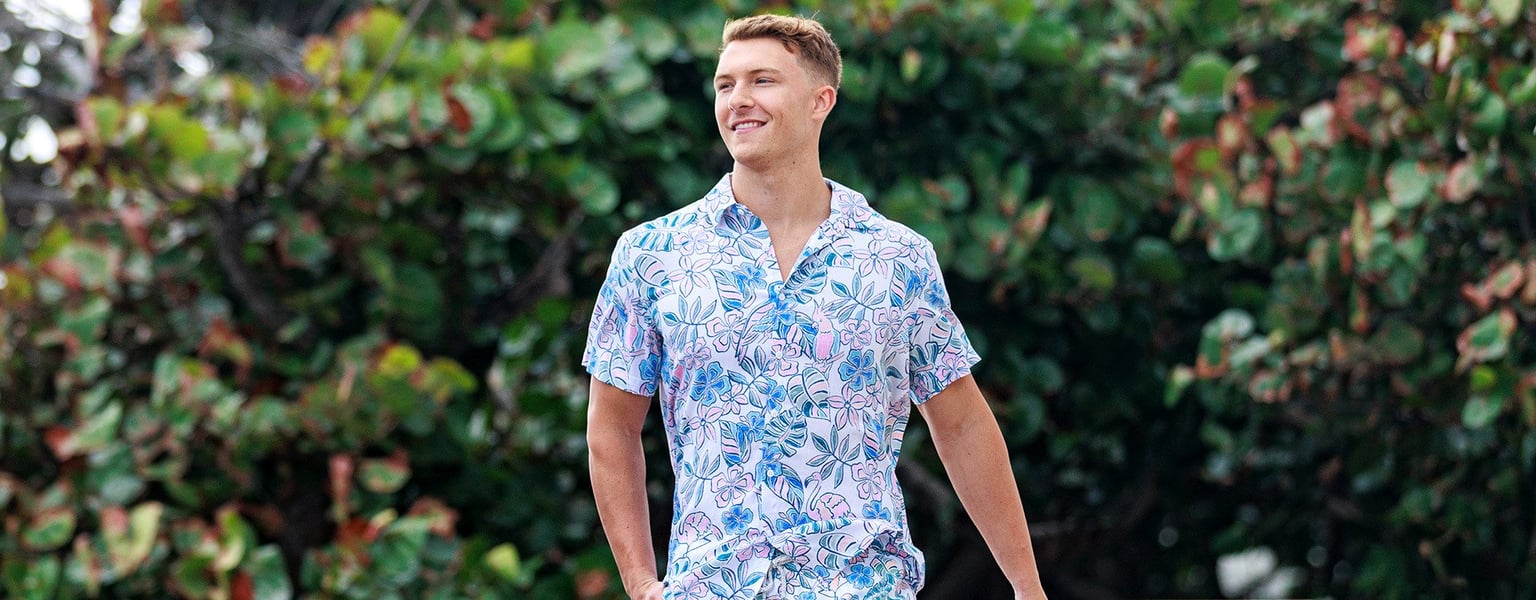 A person wearing a short-sleeve, light-colored patterned shirt is walking outdoors in front of green foliage.