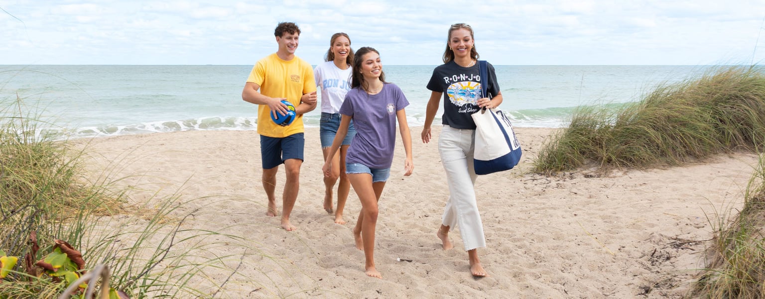 Four people walk up a sandy beach path toward grassy dunes, smiling and dressed in casual summer clothing with the ocean in the background.