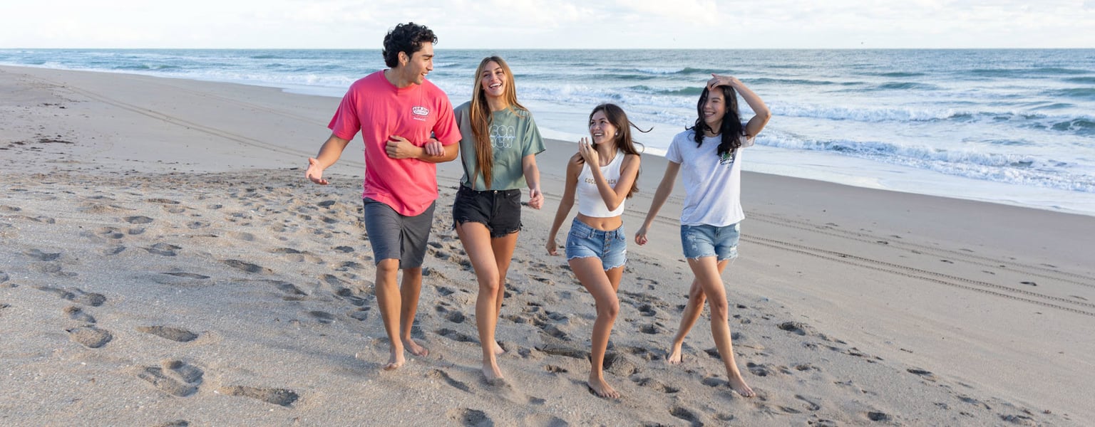 Four young adults walk barefoot together along a sandy beach near the ocean, smiling and talking on a sunny day.