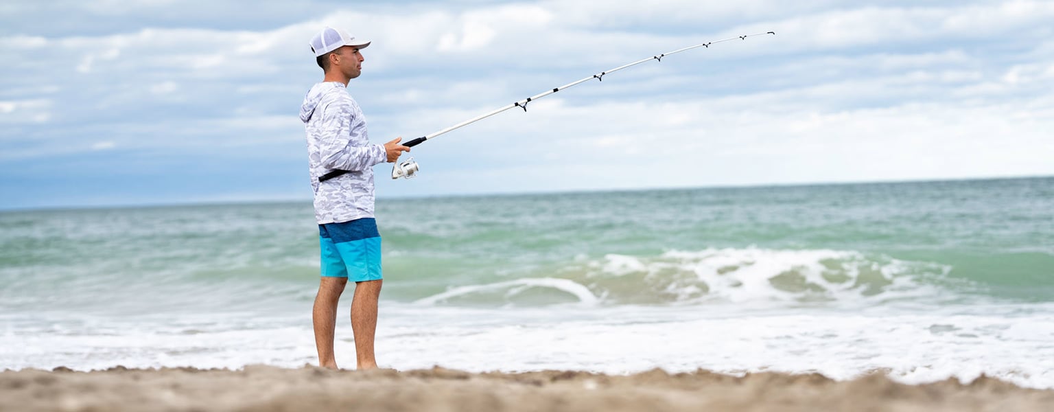 A man in a hooded shirt and hat stands on a sandy beach holding a fishing rod over the ocean.