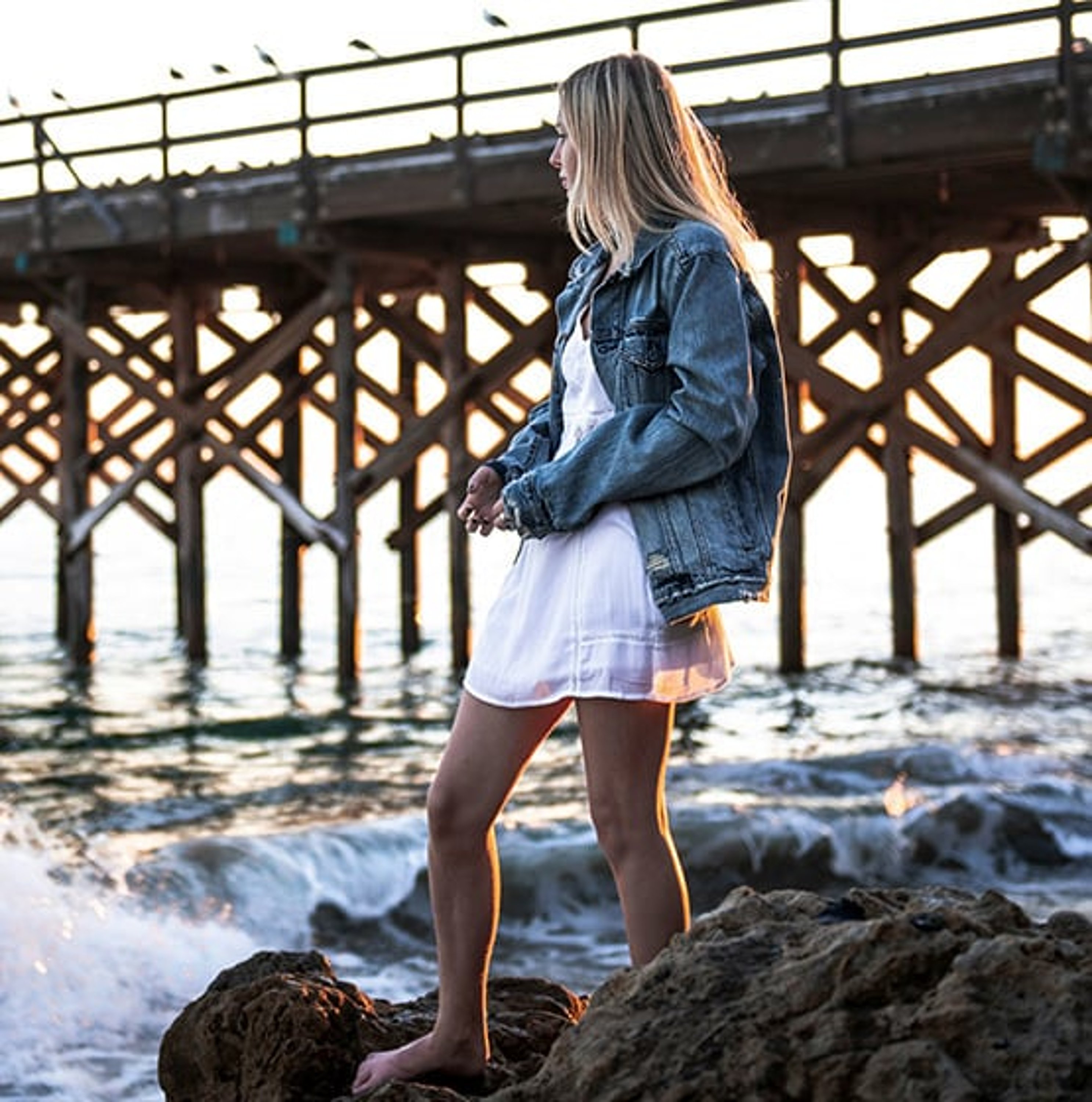 girl standing on rocks by a pier wearing a white dress and denim jacket