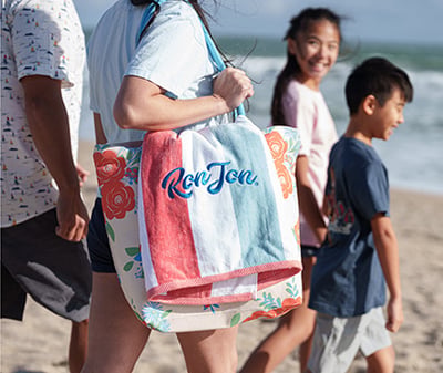 Four people walk on a sandy beach. The person in front carries a floral bag with a colorful towel that has Ron Jon written on it. The ocean and sky are visible in the background.