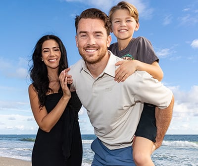 A man gives a young boy a piggyback ride while a woman stands beside them, all smiling on a beach under a partly cloudy sky.