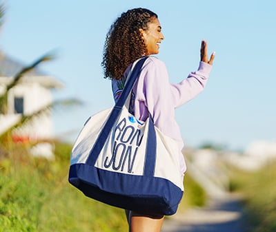 A woman with curly hair, wearing a light purple sweatshirt, carries a large blue and white bag and waves while walking outside on a sunny day.