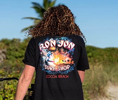 Person with curly hair wearing a black Ron Jon Surf Shop Cocoa Beach t-shirt with an astronaut surfing graphic, standing outdoors on a sunny day with greenery and a palm tree in the background.