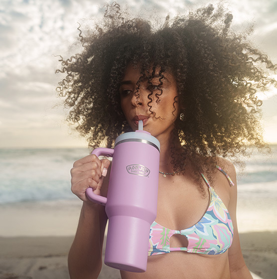 A woman in a patterned bikini drinks from a large purple insulated tumbler on a beach with the ocean in the background.