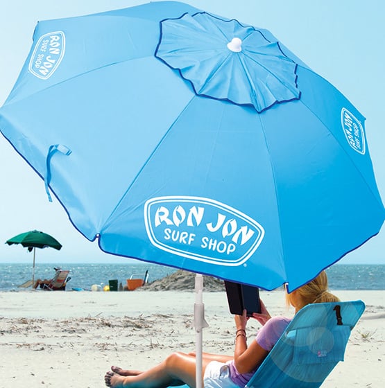 A person sits under a large blue Ron Jon Surf Shop umbrella on a sandy beach, with the ocean and another green umbrella in the background.