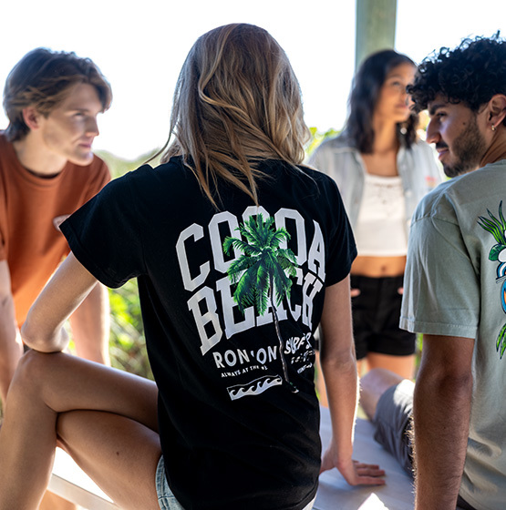Four young adults are gathered together outdoors. The person in the center wears a black t-shirt that reads “COCOA BEACH” with palm tree graphics on the back.