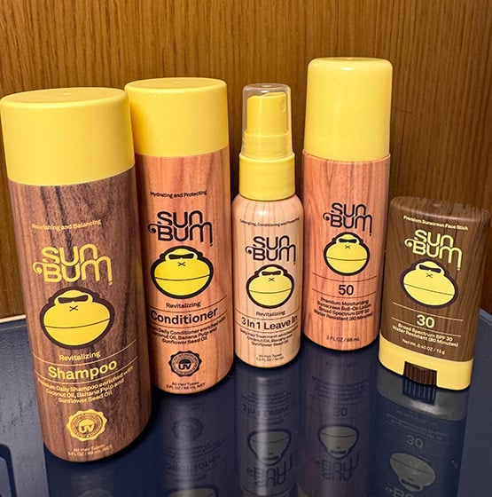 Five Sun Bum hair and skin care products, including shampoo, conditioner, leave-in spray, and sunscreens, displayed upright on a reflective surface in front of a wooden background.