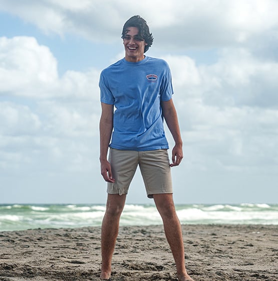 A young man stands barefoot on a sandy beach, wearing a blue t-shirt and khaki shorts, with the ocean and cloudy sky in the background.